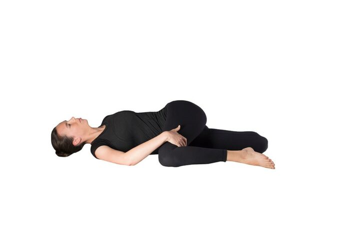 Woman doing quad stretch lying on back with left leg bent and crossed over body. Right hand is pinning down let knee, while right foot is back behind, being pulled to butt by left hand.