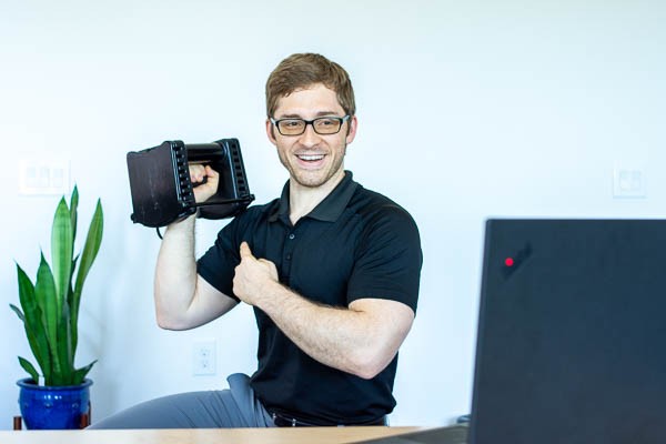 White man smiling and pointing to shoulder while holding a dumbbell and talking to a laptop