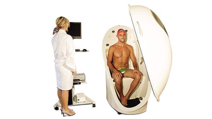 A man clothed in swim trunks and cap sitting in a BOD POD getting ready to be examined by a woman in a while coat next to a computer